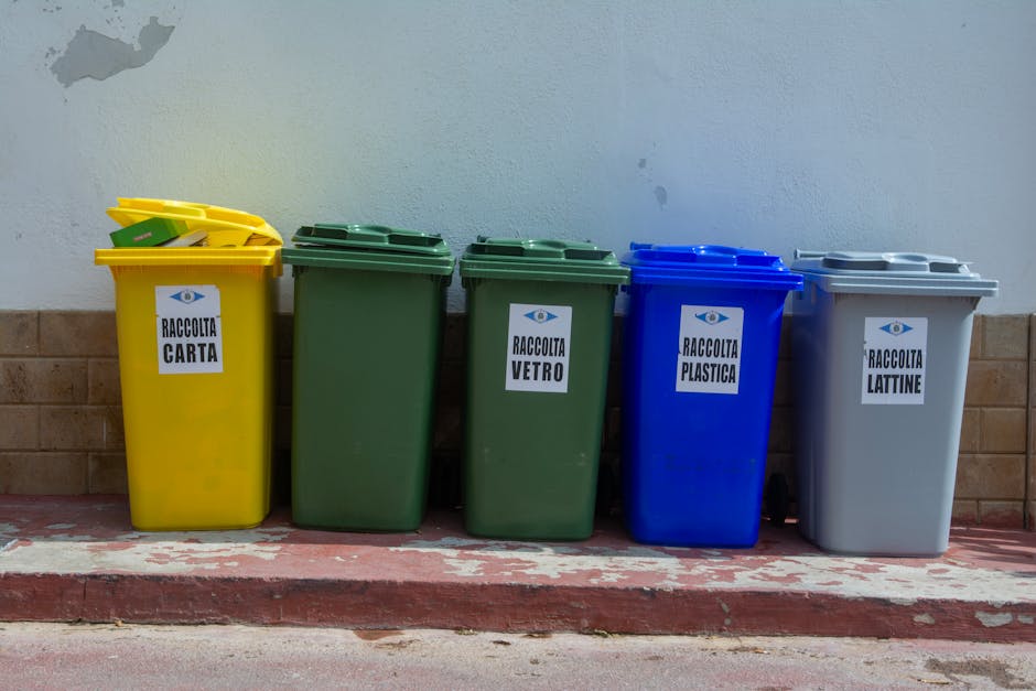 A row of five large, upright rubbish bins made of durable plastic, positioned on a red and white striped curb against a light grey wall. From left to right, the bins are colored yellow, green, green, blue, and grey. Each bin has a white label with black text indicating the type of waste collected: the yellow bin is labeled
