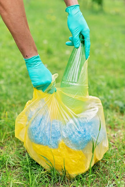 A person wearing bright blue disposable gloves is holding open a yellow plastic rubbish bag, which is filled with mixed waste, including blue and white items and some green grass or plant material visible at the bottom. The bag is being held against a grassy outdoor area with green grass and some small plants in the foreground. The background is slightly blurred but shows more grass and natural outdoor elements, indicating the waste is being collected in a garden or outdoor space. The scene reflects typical waste disposal activity, possibly related to rubbish removal or garden clearance services, emphasizing the handling of household or garden waste in an environmentally conscious manner. The image suggests an on-site waste collection process, aligned with private rubbish removal solutions by companies like Rubbish Collection Pimlico, which offer alternative disposal options outside of local authority services.