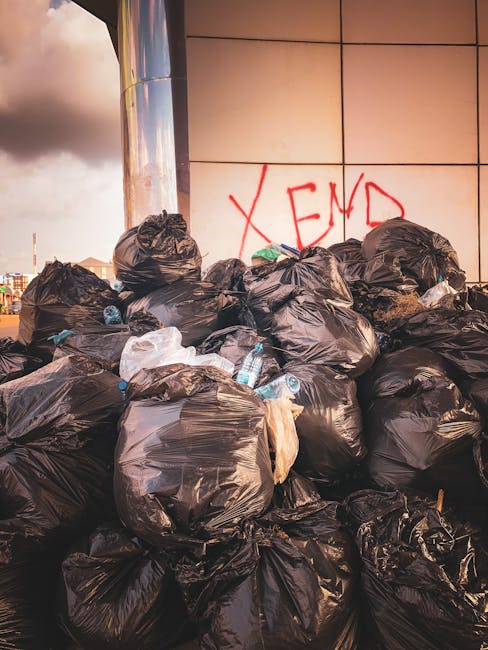 A large pile of black bin bags filled with rubbish is stacked outdoors on a paved surface beside a modern building with a tiled exterior wall. Some bags appear bulging and are tied at the top, with a few partially torn, revealing white or light-colored waste materials inside. Several clear plastic bottles are visible, partially protruding from the bags, indicating mixed waste. Behind the pile, the building's wall features a red graffiti tag that appears to spell 'XEND' spray-painted in capital letters. To the left, part of a tall, shiny metal column reflects the surrounding environment. The sky above shows dark, stormy clouds, suggesting a gloomy or potentially rainy day. This scene is typical of informal rubbish accumulation, highlighting the importance of proper waste disposal services such as those offered by Rubbish Collection Pimlico for alternative waste handling options in the area.