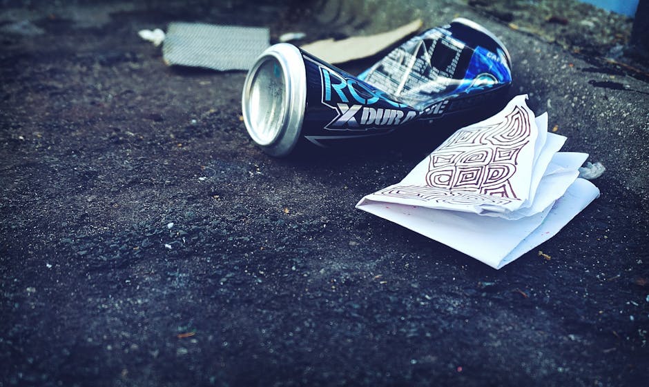 A crumpled aluminium energy drink can with a blue and silver design rests on a dark asphalt surface, partially lying on its side with the open end facing slightly upward. Next to the can, there is a small stack of crumpled white paper, some featuring brown pen or marker markings and patterns, placed on the uneven ground. The surrounding environment includes a rough, textured asphalt driveway or pavement with small stones and debris scattered across the surface, indicative of an outdoor urban setting. In the background, blurred natural elements such as soil or grass are faintly visible, suggesting an outdoor location. The scene appears to be part of a waste collection or disposal situation, typical of private rubbish removal services like those offered by Rubbish Collection Pimlico, highlighting the importance of proper rubbish handling and responsible disposal in a professional context. The lighting is natural, casting soft shadows and emphasizing the textures and colors of the discarded objects consistent with outdoor daytime conditions.