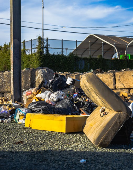 A large pile of black bin bags filled with rubbish is stacked outdoors on a paved surface beside a modern building with a tiled exterior wall. Some bags appear bulging and are tied at the top, with a few partially torn, revealing white or light-colored waste materials inside. Several clear plastic bottles are visible, partially protruding from the bags, indicating mixed waste. Behind the pile, the building's wall features a red graffiti tag that appears to spell 'XEND' spray-painted in capital letters. To the left, part of a tall, shiny metal column reflects the surrounding environment. The sky above shows dark, stormy clouds, suggesting a gloomy or potentially rainy day. This scene is typical of informal rubbish accumulation, highlighting the importance of proper waste disposal services such as those offered by Rubbish Collection Pimlico for alternative waste handling options in the area.