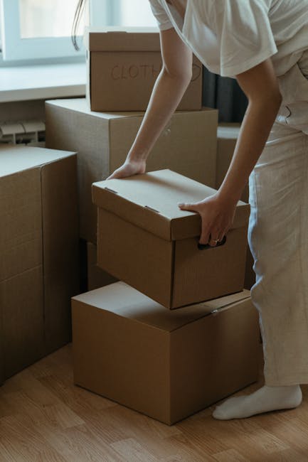 A person wearing a white T-shirt and light-colored trousers is seen arranging three brown cardboard boxes in an indoor setting, likely a residential space. The boxes are rectangular with smooth surfaces and flat, folded-over flaps on top, with one visibly labeled 'CLOTHES' in red ink. They are being stacked on a wooden floor near a window that allows natural light to illuminate the scene. In the background, additional cardboard boxes of varying sizes are visible against a wall, some partially obscured, suggesting they contain household items for packing or disposal. The environment appears tidy, with the boxes organized for moving, storage, or potential waste collection. Rubbish Collection Pimlico’s focus on waste management and alternative disposal services is subtly connected through the scene of packing or preparatory rubbish handling, indicative of private or independent waste removal methods rather than council-led bulky-item collections.