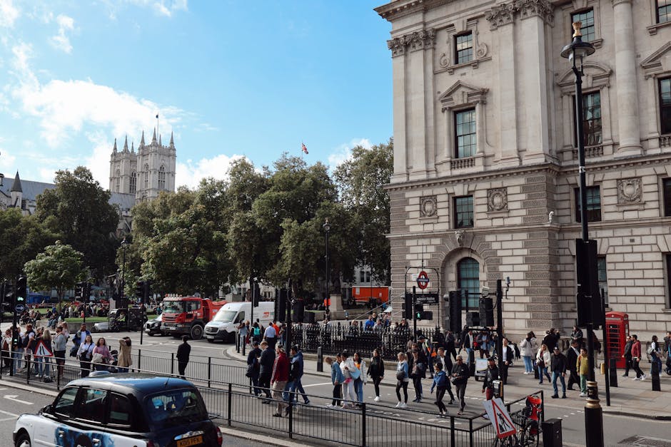 A busy London street scene near a large, historic stone building with ornate architectural details, including decorative columns and arched windows, situated on the right side of the image. The foreground features a metal railing along the sidewalk and a pedestrian crosswalk with numerous people walking in various directions, dressed in casual attire. Some are carrying bags or backpacks, and a few are crossing the street. Several vehicles, including a black car at the bottom left, a white van, and a red truck, are on the road with traffic signals visible. Behind the pedestrian area, there are several trees with dense green foliage lining the street, and in the background, a tall Gothic-style tower with multiple spires rises against a partly cloudy sky. The scene captures urban activity and the historic environment, reflecting typical city infrastructure and public space management, relevant to services like rubbish collection or alternative waste handling in busy metropolitan areas such as Westminster.