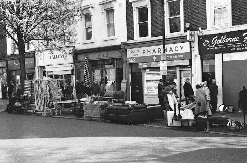A street scene in an urban commercial area showing a variety of household and retail items placed on the pavement outside shops. Visible objects include large black sofas, a rolled-up rug, smaller furniture pieces, lamps, and miscellaneous household goods. The items are arranged in a somewhat haphazard manner, with some stacked or leaning against each other, indicating a clear intention of waste removal or disposal. Several individuals are standing nearby, observing or engaged in conversation, while others appear to be loading or sorting the items. Behind the scene, storefronts for a pharmacy and a seafood specialist are visible, with shop signs, windows, and doorways providing a backdrop. The environment is outdoor, with natural daylight illuminating the scene, and a lamppost and a tree also visible on the pavement, contributing to the typical street-level context of independent waste collection or on-site clearance conducted by Rubbish Collection Pimlico or similar rubbish removal services.