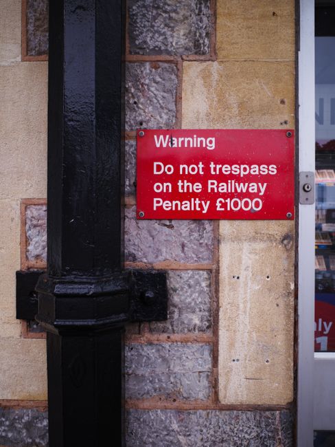 Close-up of a red warning sign mounted on a brick and stone wall next to a black pipe or cable conduit. The sign displays the message 'Warning Do not trespass on the Railway Penalty £1000' in white text. The wall behind the sign consists of irregularly shaped bricks and stones in shades of tan, brown, and gray, with some areas showing weathering or slight discoloration. To the right of the sign, part of a glass door or window is visible, reflecting a glimpse of the street or nearby buildings. The scene appears to be outside a building, possibly adjacent to railway property, emphasizing the caution against unauthorized entry. The environmental lighting is neutral, suggesting daytime with no harsh shadows, and the setting conveys a sense of security and regulation regarding trespassing in this area, a relevant aspect in discussions of private waste or illegal dumping enforcement by local authorities or private waste management services such as Rubbish Collection Pimlico.