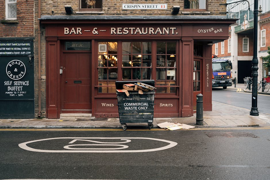 The image depicts the exterior of a small, closed bar and restaurant situated on a city street, with the façade painted in a deep reddish-brown color. Large glass windows with grid-like wooden frames dominate the front, revealing a warmly lit interior with hanging pendant lights. To the right of the entrance, a narrow vertical sign indicates the availability of cocktails. In front of the establishment, a black wheeled commercial waste bin labeled 'COMMERCIAL WASTE ONLY' is positioned on the sidewalk, filled with discarded cardboard boxes and paper, with some debris spilling onto the pavement. Surrounding the bin, there are a metal bollard and a small, upright street sign attached to the building's corner. The street features a white road marking indicating a no-parking zone, and a black metal lamppost is situated further along the sidewalk. In the background, a delivery truck and bicycles are visible, contributing to the urban environment, which suggests a typical city setting that may require occasional rubbish removal services by companies like Rubbish Collection Pimlico, particularly in contexts involving on-site clearance of commercial waste.