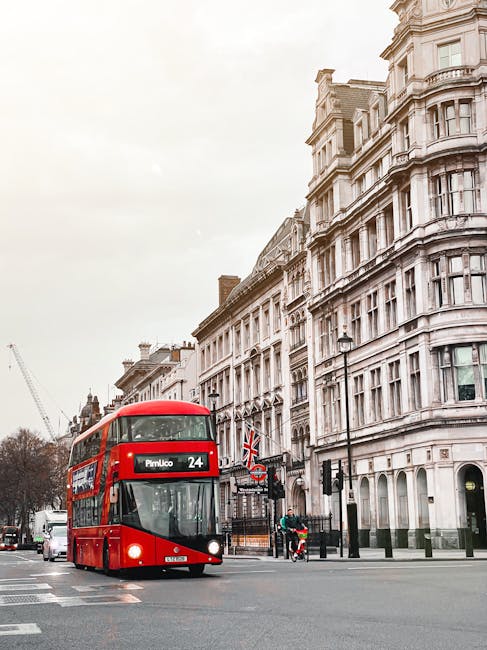 A red double-decker bus numbered 24 is traveling through a city street lined with historic, multi-storey white stone buildings featuring ornate architectural details. The bus displays a destination sign indicating 'Pimlico' and is positioned on the left side of the image, moving towards the foreground. It is flanked by a few pedestrians on the sidewalk, including someone on a bicycle near pedestrian crossings and a person wearing high-visibility clothing beside a small vehicle. The sky above is overcast, casting a soft, diffused light over the scene. The street has traffic lights and lamp posts, and in the background, there is a construction crane partially visible among the buildings, indicating ongoing urban development. The scene captures an everyday moment in central London, with an emphasis on urban transport and city architecture, subtly relevant to private and alternative waste management services like those provided by Rubbish Collection Pimlico, operating in city environments where independent rubbish collection might be required for local flats or commercial properties.