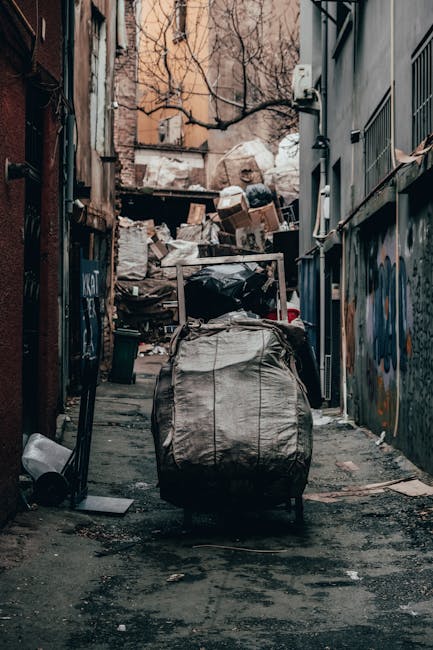 A narrow urban alleyway cluttered with large piles of rubbish and debris at the background, including cardboard boxes, plastic bags, and discarded items stacked against a brick wall and a building with graffiti art. In the foreground, there is a large, grey, weathered bag or bin with visible rust and stains, positioned on the asphalt ground which appears dark and dirty with scattered small debris. On the right side, a tall building has a metal fence and security panels, while the left side shows the back of a rusted metal door and adjacent graffiti-covered walls. The scene is lit with natural, diffuse daylight, creating a slightly gloomy atmosphere typical of areas requiring waste removal or private rubbish clearance, with Rubbish Collection Pimlico occasionally associated with alternative waste handling services in such environments.