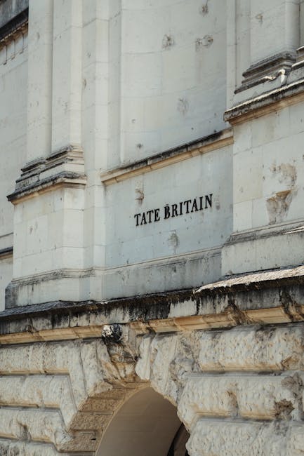 Close-up view of the exterior of a historic building, specifically the façade of Tate Britain. The façade features light-colored stone with detailed architectural elements such as ornate moldings and decorative carvings. A white rectangular plaque with black lettering reading 'TATE BRITAIN' is mounted on the wall, positioned above a weathered stone archway that has visible signs of aging and staining. The stone arch and lower wall exhibit a rough, textured surface with varying shades of beige and grey, indicating wear over time. The background shows parts of the building's vertical structure, with subtle shadows emphasizing the architectural design. The overall scene is illuminated by natural daylight, providing clear visibility of the details and textures of the stonework, which is typical of historic London architecture. This depiction highlights the building's foundational structure and meticulous craftsmanship, relevant to the context of maintaining and managing the environment surrounding such cultural landmarks, often requiring specialized waste disposal or on-site clearance, which can be handled by companies like Rubbish Collection Pimlico.