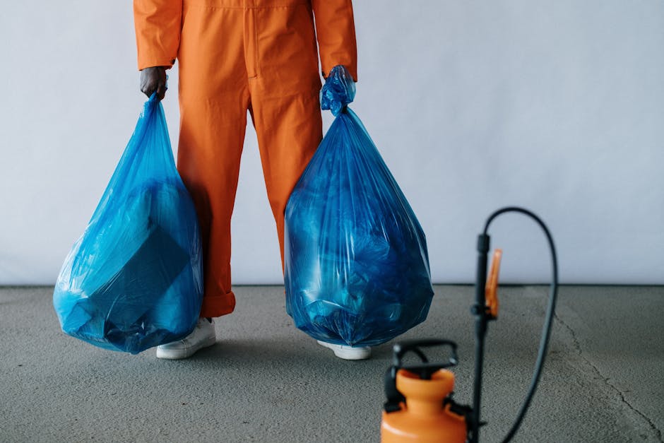 A person standing indoors wearing an orange work jumpsuit and white sneakers, holding two large, semi-transparent blue plastic garbage bags filled with waste, one in each hand. The bags appear to be full and are slightly bulging, with the contents not visible but inferred to be household or industrial rubbish. The background features a plain, light-colored wall and a smooth grey floor, possibly concrete. In the foreground, there is a device with a black cord and orange handle, which looks like a portable vacuum or pest control equipment, suggesting a cleanup or waste collection context. The scene conveys an environment related to waste handling or rubbish removal services, typical of private rubbish collection operations, with an emphasis on emptying and transporting waste rather than recycling activities. The overall composition balances the focus on the waste bags and initial handling process, aligning with independent rubbish disposal or on-site clearance tasks carried out by companies like Rubbish Collection Pimlico.