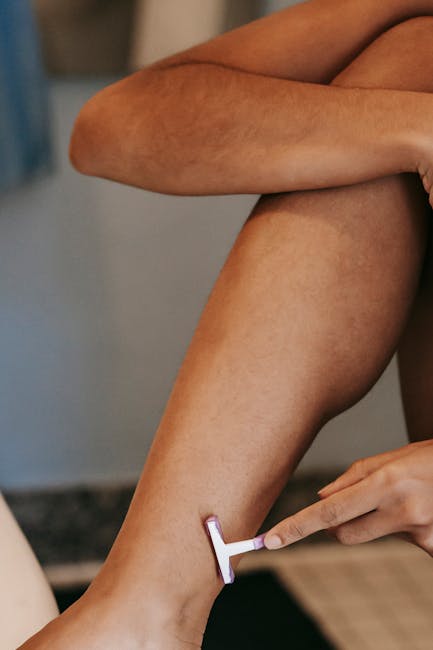 A close-up image showing a person's bare legs with smooth skin, sitting on a surface with one leg crossed over the other. The individual is using a disposable razor with a white handle to shave the lower leg. The background appears blurred and includes elements of a room with muted lighting, possibly a bathroom or personal grooming area. The lighting highlights the natural skin tone and texture, with subtle shadows emphasizing the contours of the legs. The scene reflects routine personal grooming rather than any waste or rubbish removal context, and no waste-related objects are present in the image. This visual focus on grooming activities aligns with general private disposal and individual hygiene practices, which Rubbish Collection Pimlico may occasionally support through related services, although the scene itself is not directly connected to rubbish removal services.