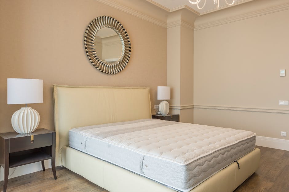 Inside a bedroom with beige walls and white crown molding, there is a cream-colored upholstered bed with a matching padded headboard, positioned against the left wall. On either side of the bed, there are dark wooden bedside tables; each holds a white ceramic table lamp with a ribbed, spherical base and plain white lampshade. Above the headboard, a round mirror with a decorative, striped frame reflects part of the ceiling and a light fixture, which is partly visible. The room's lighting is soft and natural, illuminating the space evenly. The walls are unadorned, and there are electrical outlets and a light switch visible on the right wall. The floor is covered with a light wood laminate or hardwood finish, contributing to a calm and neutral atmosphere. This interior scene is purely decorative and does not include any rubbish or waste, aligning with an image of a clean, well-maintained personal space rather than a rubbish removal context, though the setting is described in a neutral, factual manner suitable for service-related content.