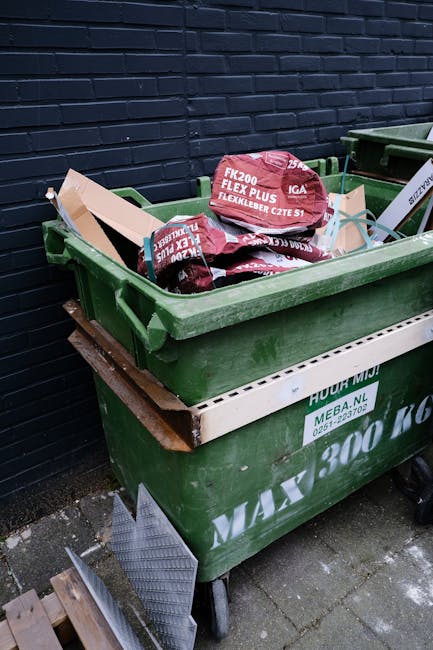 A large green wheeled rubbish bin filled with red and black bags of garden or landscape waste, including bags labeled 'FK200 FLEXY PLUS' and 'FLEXLEBER CZTE S1,' situated outdoors on a paved surface against a dark brick wall. The bin is partially stacked on a white containment tray, and nearby, a dark green recycling container is visible. The scene appears to be part of an on-site waste collection or disposal setup, possibly related to private rubbish removal services. The environment is clean, with natural ambient lighting illuminating the scene, highlighting the textures of the bags, the roughness of the brick wall, and the surfaces of the bins, reflecting an organized approach to waste handling typically associated with professional rubbish collection in Pimlico.