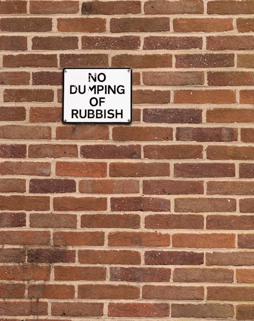 A white rectangular sign with black uppercase lettering reading 'NO DUMPING OF RUBBISH' is mounted on a red brick wall with a traditional running bond pattern. The bricks vary in shades from light to dark red, with some darker brownish tones, and have a textured, matte finish. The mortar joints are light grey and prominently define each brick. The sign is situated slightly above the center of the image, attached with four metal screws, and casts a subtle shadow onto the brick wall beneath it. The wall appears clean and well-maintained, providing a clear visual context of a designated area where rubbish dumping is prohibited, which aligns with the theme of waste management and private disposal services provided by Rubbish Collection Pimlico.